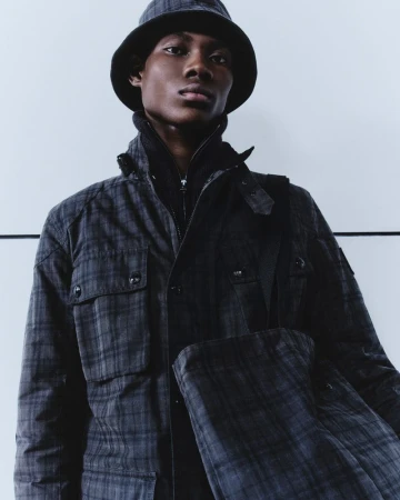 Dark-skinned male model in patterned jacket and bucket hat poses against a white wall, showcasing fashion in a studio setting.