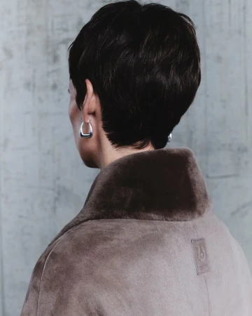 Model seen from behind wearing a brown shearling collar coat with short dark hair and silver earrings against a textured grey wall