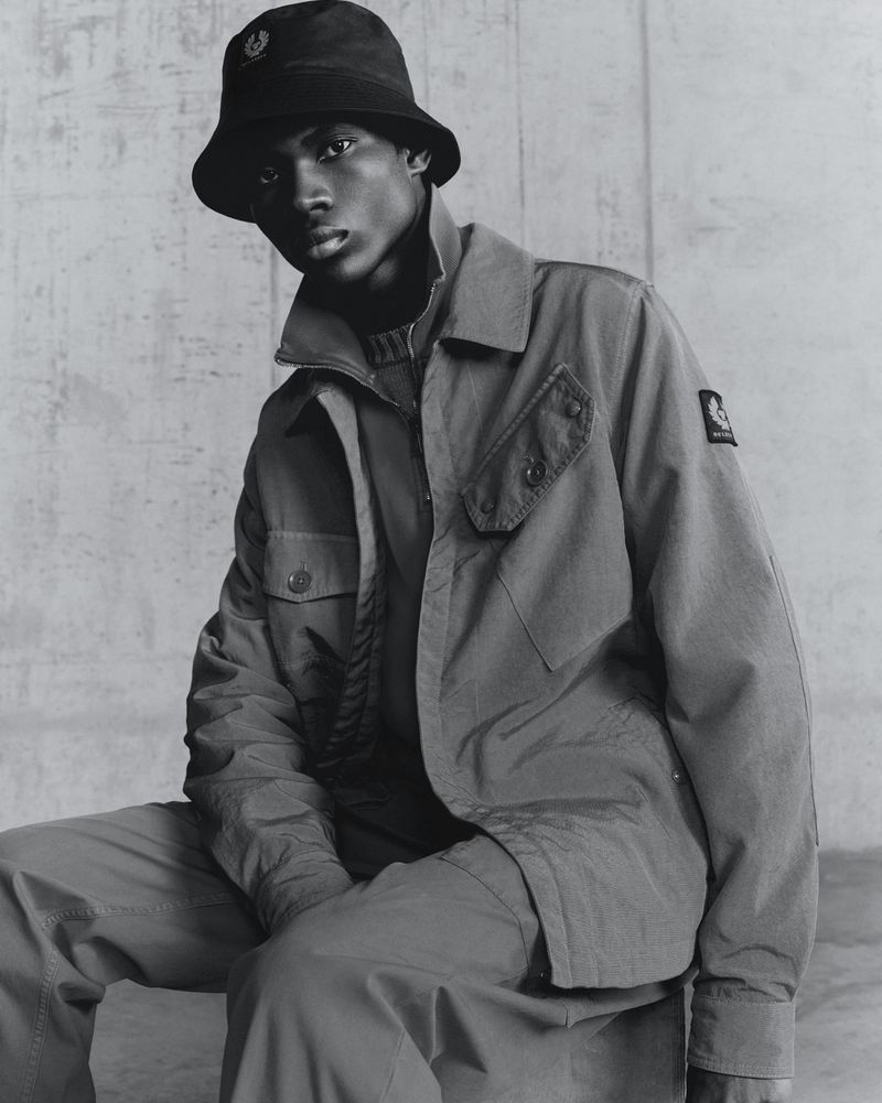 Portrait of an African American male fashion model in a bucket hat and utility jacket sitting in front of a textured wall.