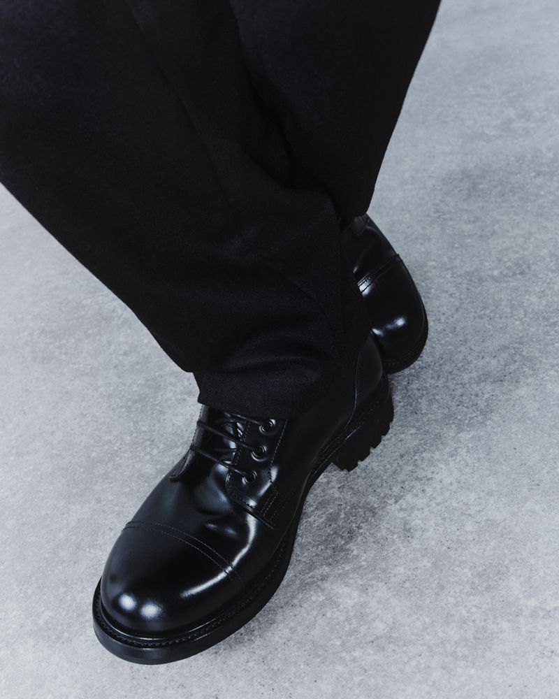 Close-up of black leather lace-up dress shoes on a grey concrete surface, paired with dark trousers, showcasing footwear fashion.