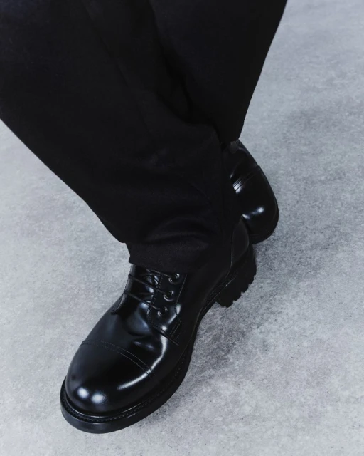 Close-up of black leather lace-up dress shoes on a grey concrete surface, paired with dark trousers, showcasing footwear fashion.