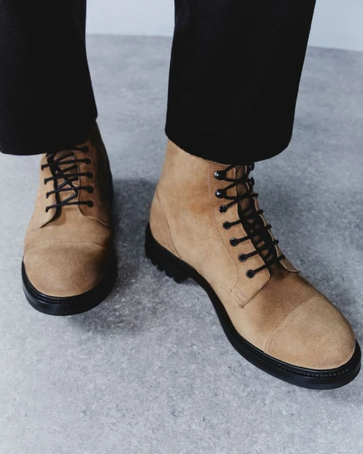 Close-up of tan suede lace-up boots with black soles and laces, styled with black trousers against a mottled grey backdrop.