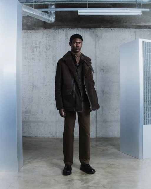 A man wearing a brown jacket and trousers stands in an industrial style room with concrete walls and metal accents for a fashion shot.