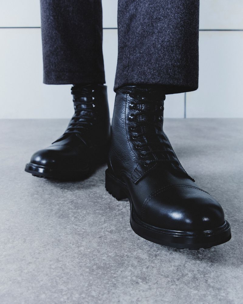 Black leather lace-up boots are worn with grey wool trousers against a bright grey backdrop. The footwear dominates the shot.