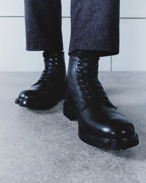 Black leather lace-up boots are worn with grey wool trousers against a bright grey backdrop. The footwear dominates the shot.