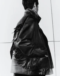 Model wearing a black fringed leather jacket against a light panelled wall in a Belstaff autumn/winter lookbook image.