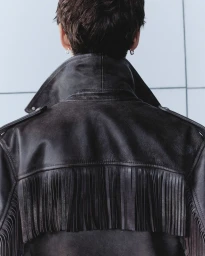 Rear view of a person wearing a black fringed leather jacket against a light tiled wall.