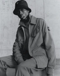 Black man wearing bucket hat and utility jacket seated against plain concrete background for Belstaff AW/24