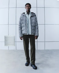 Model standing against a tiled exterior wall wearing a grey puffer jacket, light shirt, dark trousers, and black shoes.