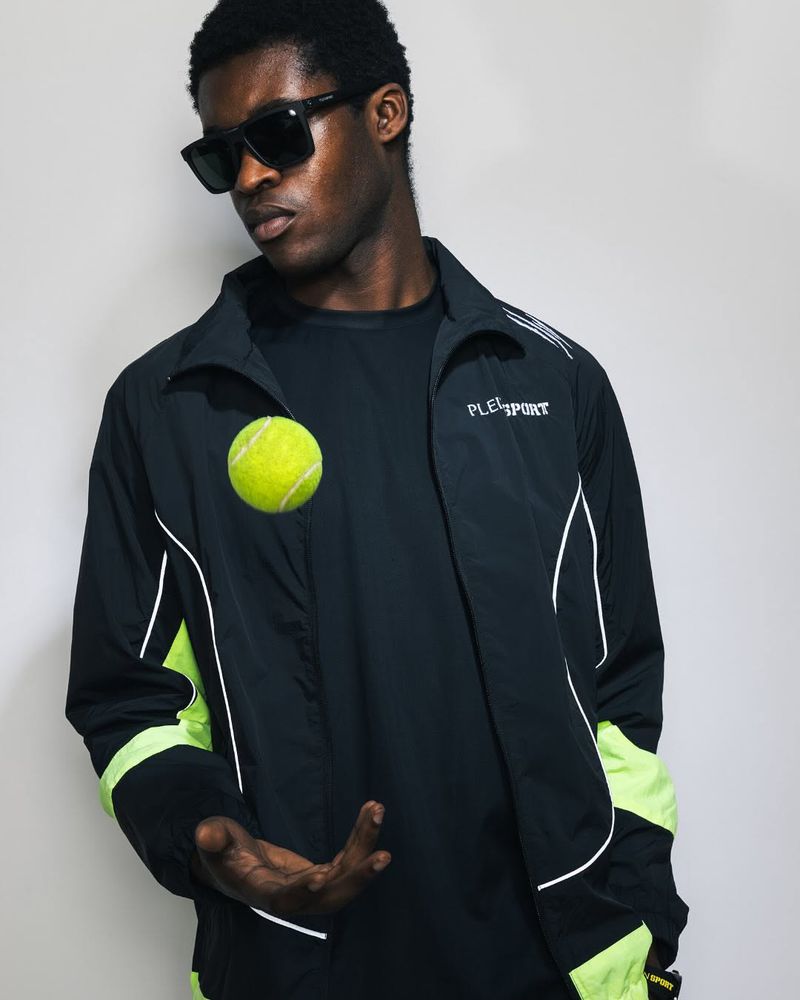 Young Black male model in a black and neon green sport jacket poses with a tennis ball in a studio against a light grey background.
