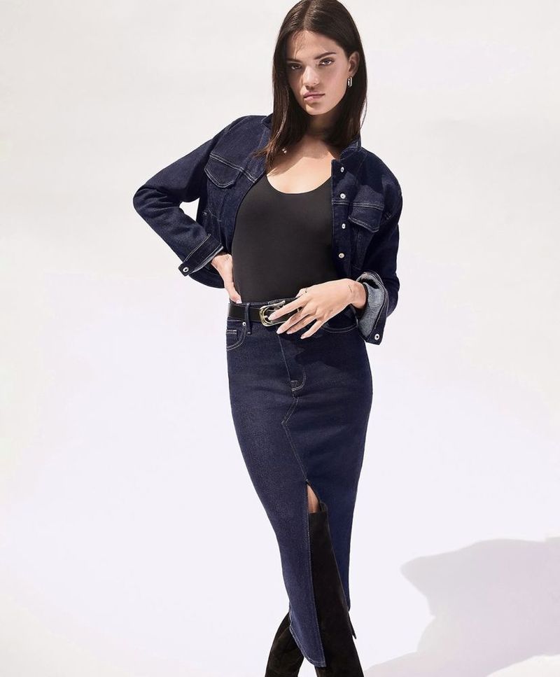 Brunette woman posing in a denim jacket, black top, denim skirt, and boots in a studio setting with a bright, white background.
