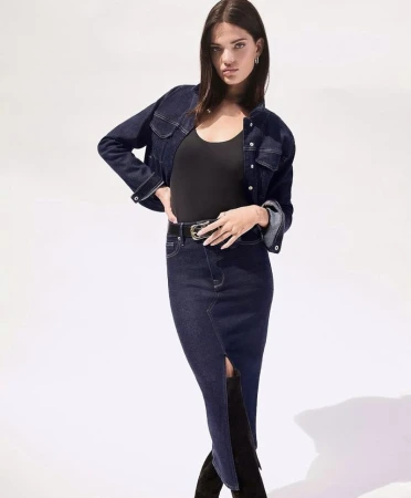 Brunette woman posing in a denim jacket, black top, denim skirt, and boots in a studio setting with a bright, white background.