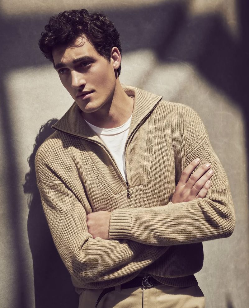An adult male with dark hair poses indoors, wearing a beige sweater and khaki pants, standing against a wall with a neutral background.