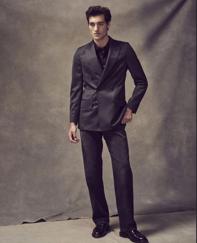 A fashionable adult male stands in a photo studio wearing a suit, a button-down shirt, and leather shoes in a full-length studio shot.