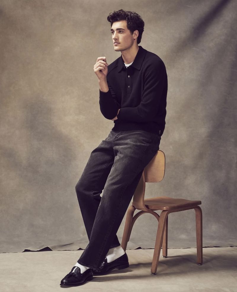 A young adult male sits on a wooden chair wearing a black long-sleeved polo shirt, black pants, white socks, and black loafers in a studio.