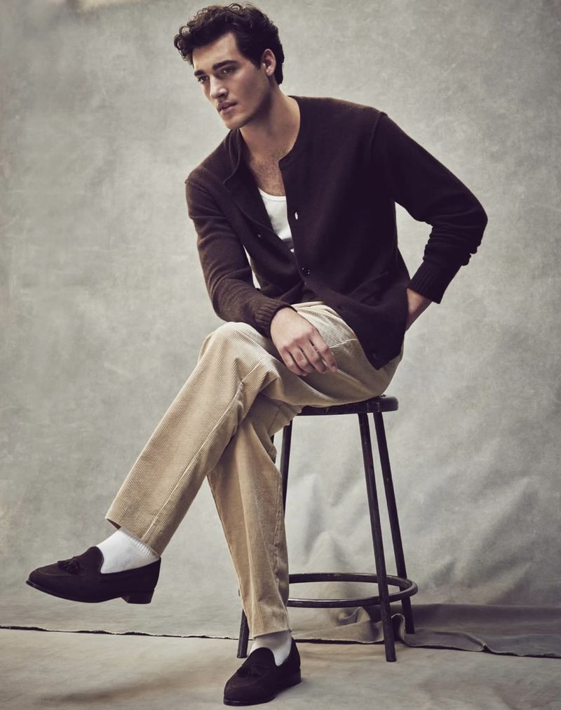 Man sitting on a stool in a studio, wearing a brown cardigan, white t-shirt, beige corduroy pants, white socks, and brown loafers.