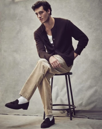 Man sitting on a stool in a studio, wearing a brown cardigan, white t-shirt, beige corduroy pants, white socks, and brown loafers.