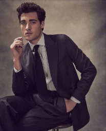 Model Adam Sattrup in a dark tailored suit, striped collared shirt, and black tie seated against a textured backdrop.