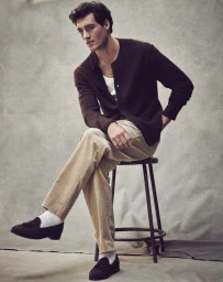Model seated on a stool wearing a dark brown cardigan, beige trousers, white socks, and dark loafers against a textured grey backdrop.