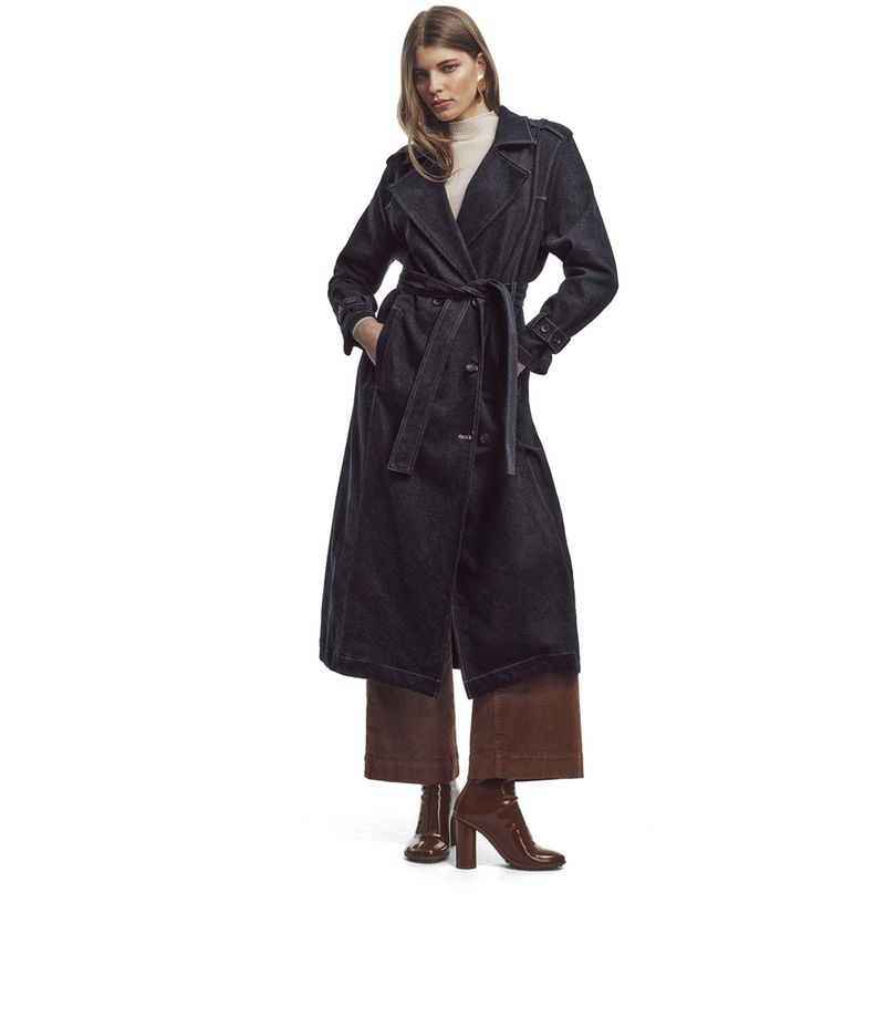 A woman stands wearing a long, dark-blue belted denim trench coat, brown pants, and heeled boots against a bright white background.