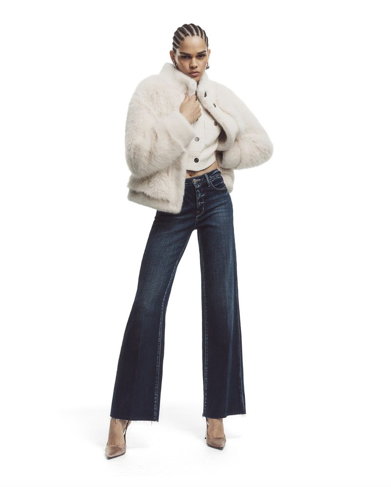 Fashionable adult female poses in a light faux fur coat, dark flared jeans, and heels against a white backdrop in a studio shot.
