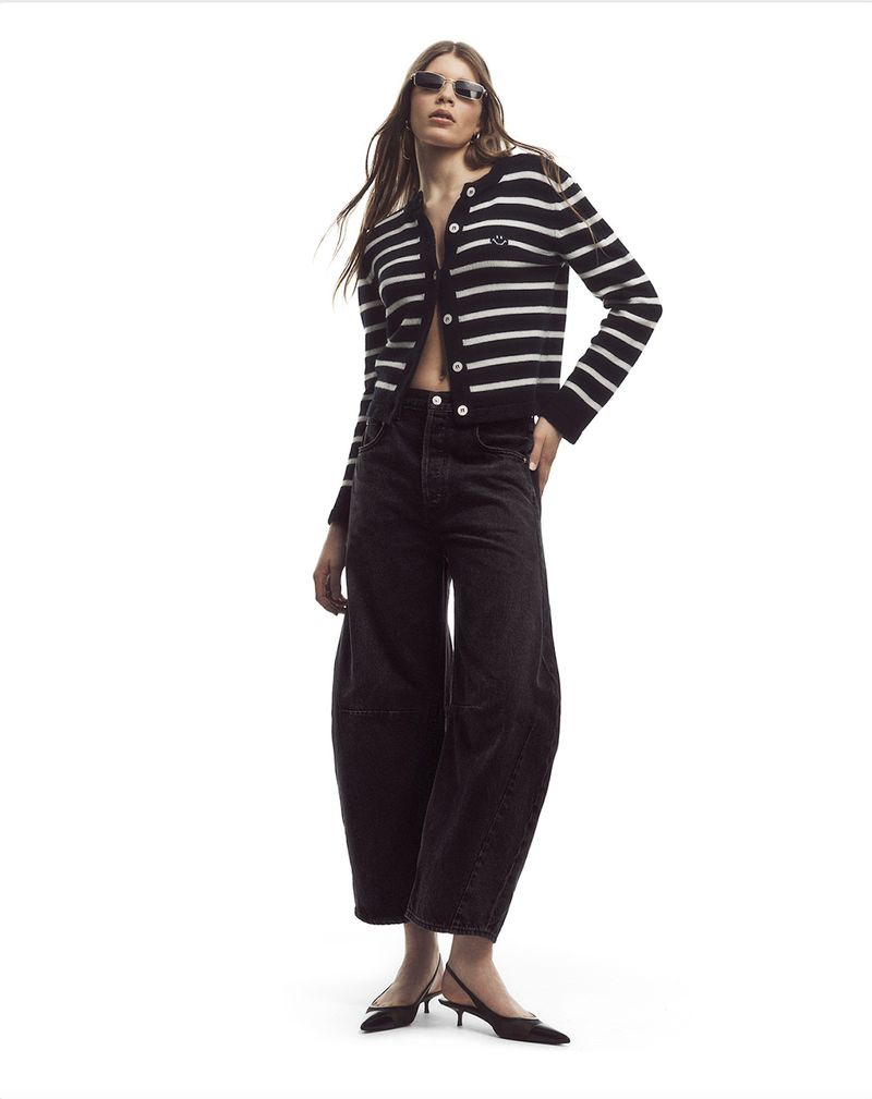 A young adult woman in a black and white striped cardigan, loose dark jeans, and slingback heels, poses in a full-body studio shot.
