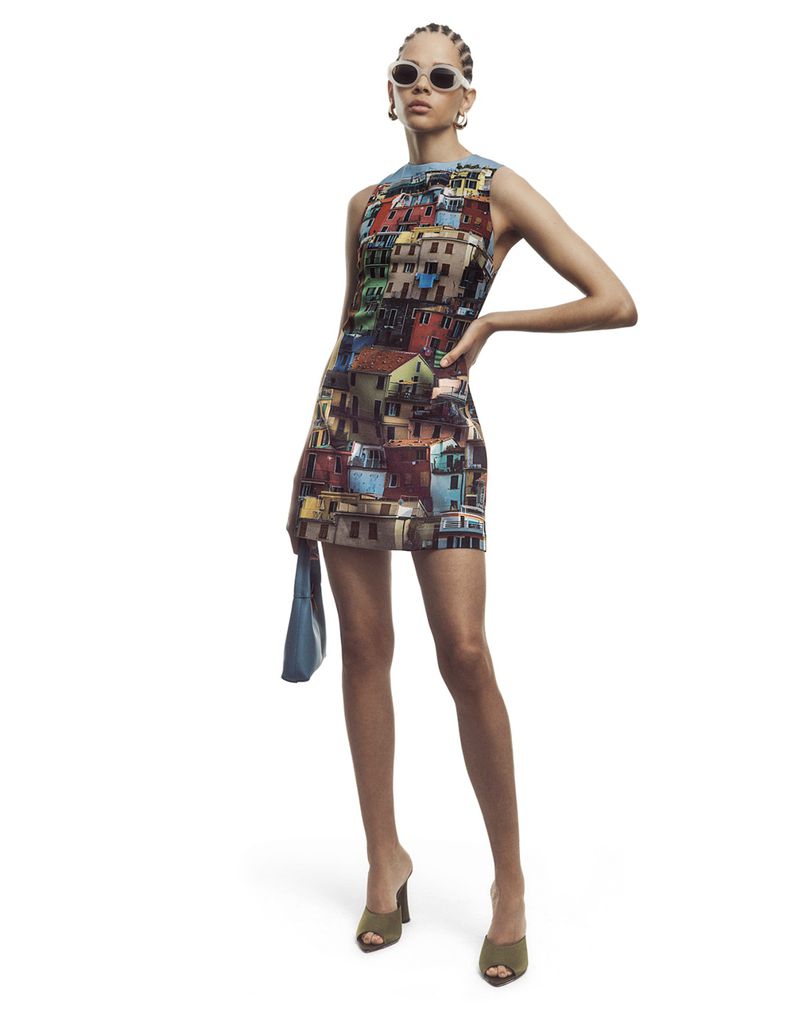 A woman in a mini dress with a building pattern poses with sunglasses and a handbag against a white backdrop in a studio shot.