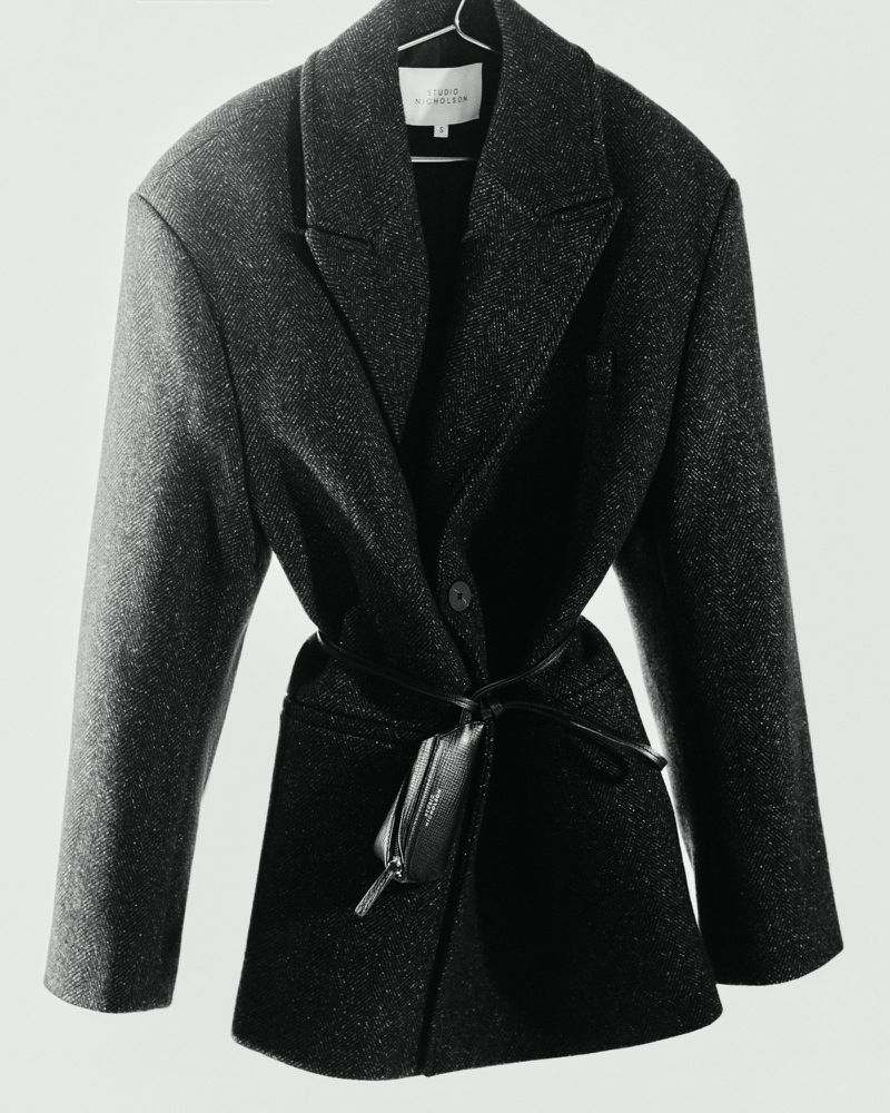 Dark grey wool coat with a leather belt detail draped on a hanger, set against a plain white background in a studio setup.