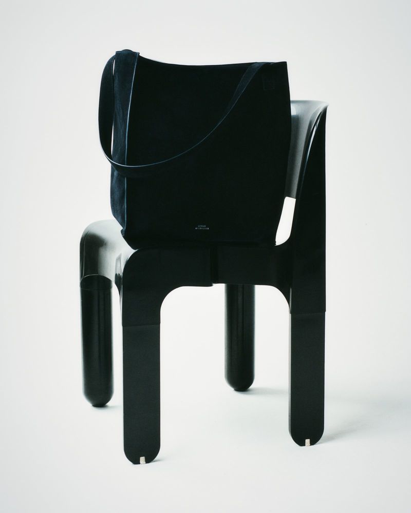 Black suede tote bag on a black plastic chair against a white seamless background in a studio product shot for fashion.