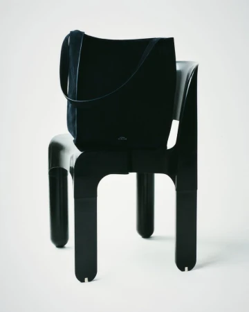 Black suede tote bag on a black plastic chair against a white seamless background in a studio product shot for fashion.