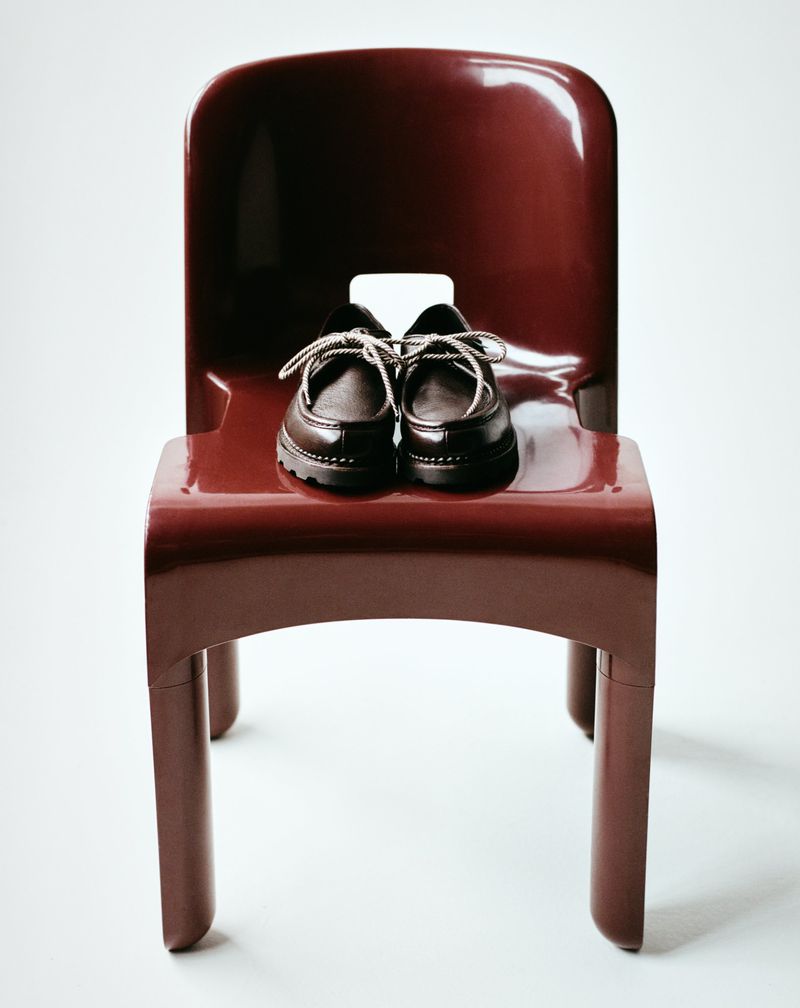 A pair of black leather baby shoes sits atop a glossy red plastic chair against a bright white backdrop, studio-lit with soft shadows.