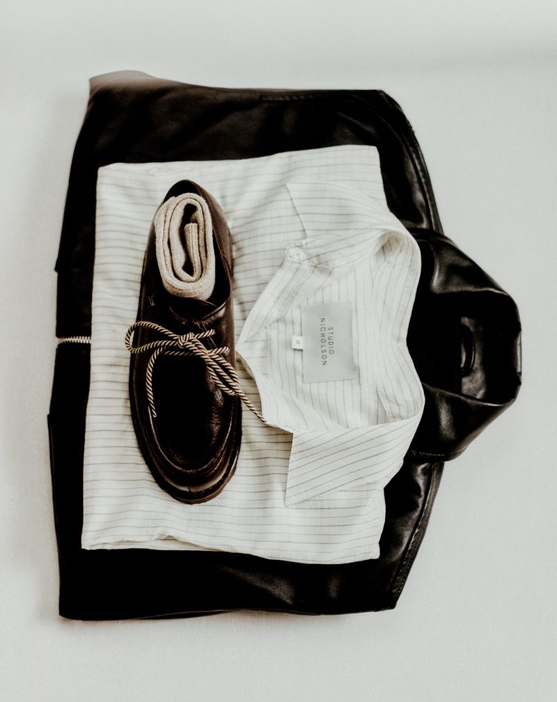 Flat lay of a brown leather shoe with a rolled sock, a striped shirt, and a black leather jacket on a white surface.