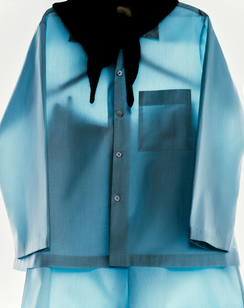 Light blue collared shirt with buttons and a pocket, styled with a black bandana, hangs on a hanger against a white backdrop.