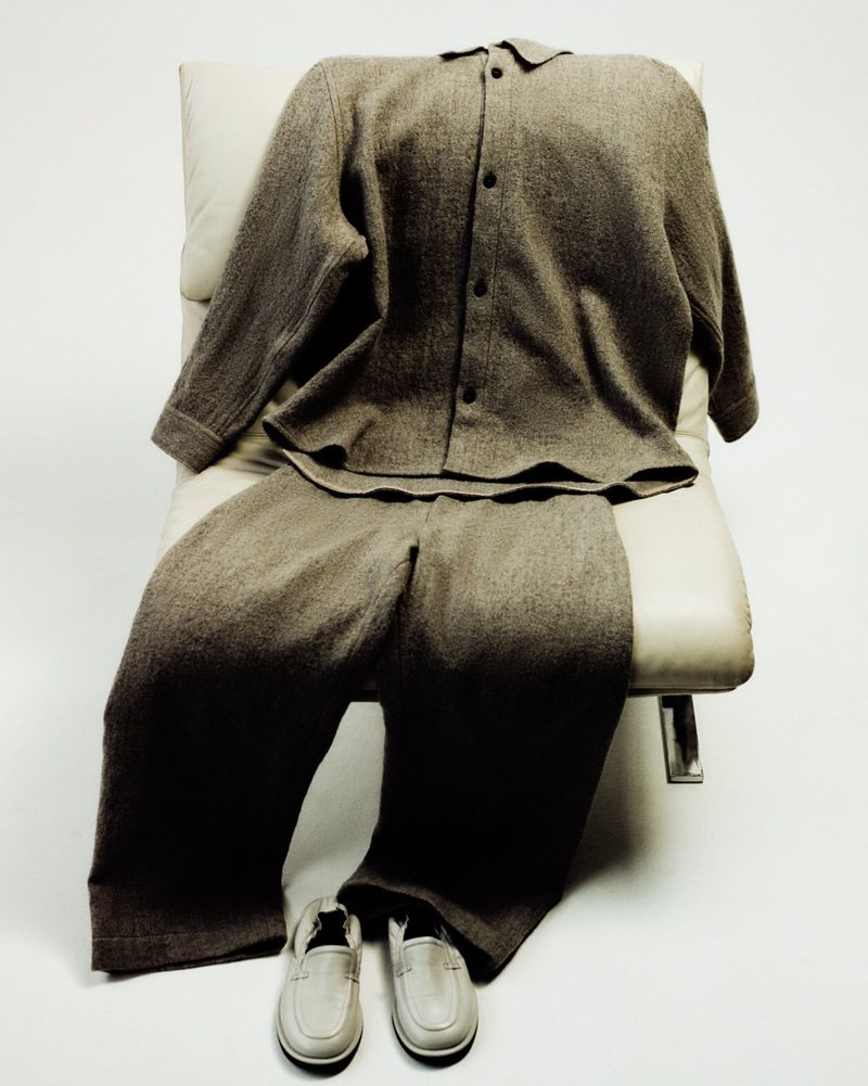 Neutral-toned lounge wear set including long-sleeve buttoned shirt, pants, and shoes, arranged on a modern chair in a bright studio.