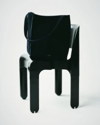 Black suede tote bag resting on a modern glossy black sculptural chair against a white background.