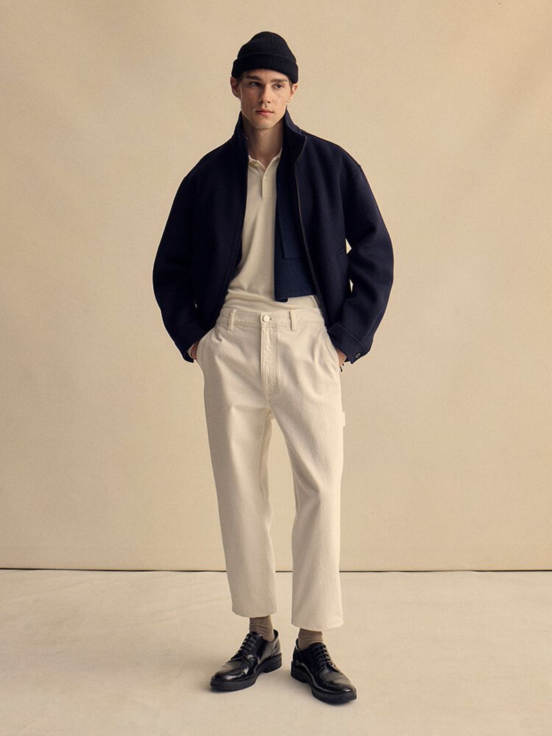 Full-length shot of an adult male wearing a navy jacket, cream polo shirt, white trousers, black beanie, and black dress shoes.