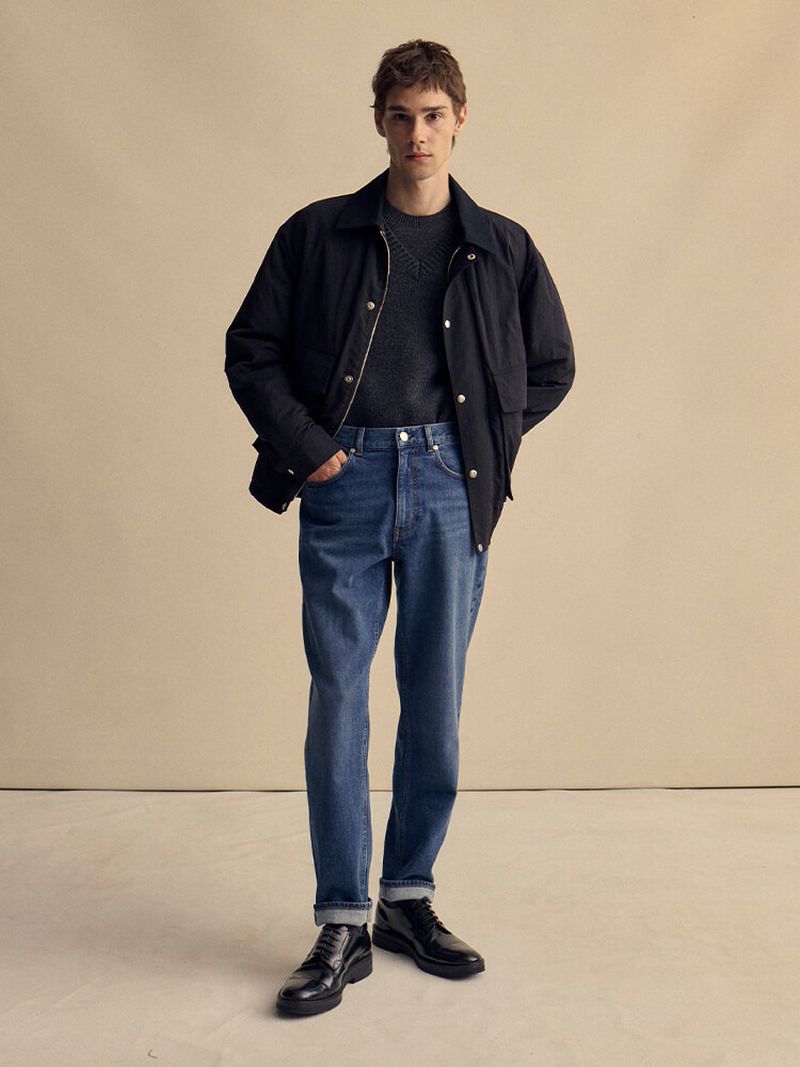 A full-length shot of a male fashion model in blue jeans, a dark sweater and jacket, and black dress shoes, standing in a neutral-toned studio.