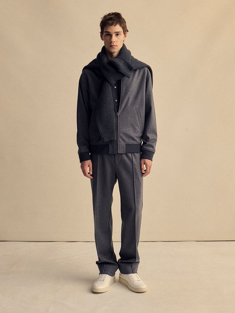 Full-length shot of an adult male wearing a grey bomber jacket, charcoal scarf, and grey pants in a neutral indoor studio setting.