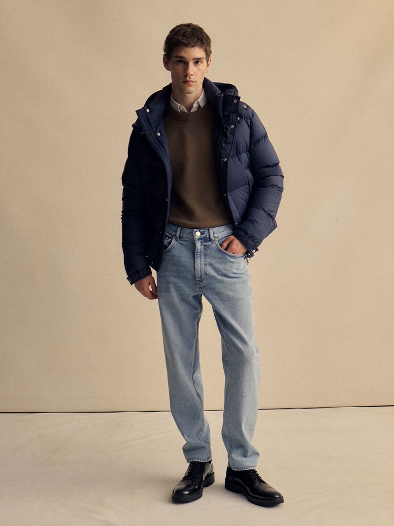 Model Patryk Lawry posing in a navy puffer jacket, brown knit sweater, light wash jeans, and black shoes against a beige background.
