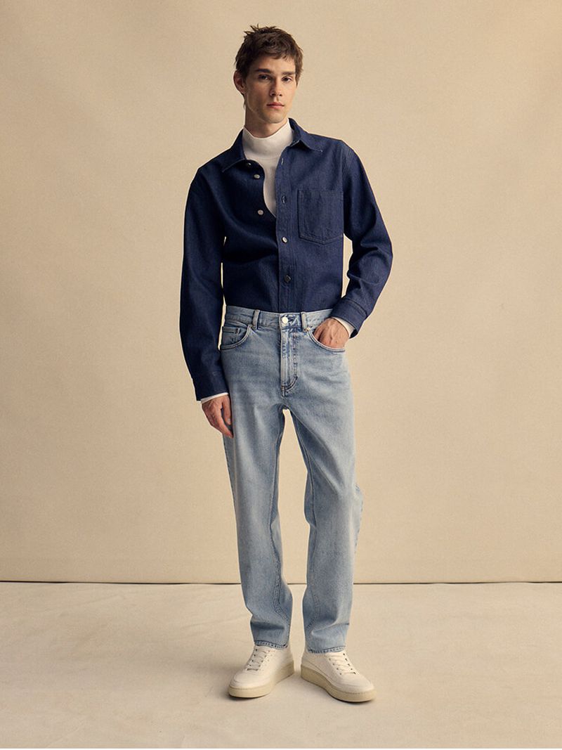 A full-length studio shot of a male model in a navy denim jacket, white turtleneck, light blue jeans, and cream sneakers against beige backdrop.