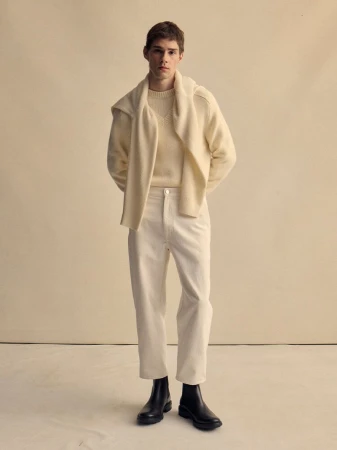Model Patryk Lawry standing in a cream knit sweater, matching trousers, and black boots against a neutral beige backdrop.