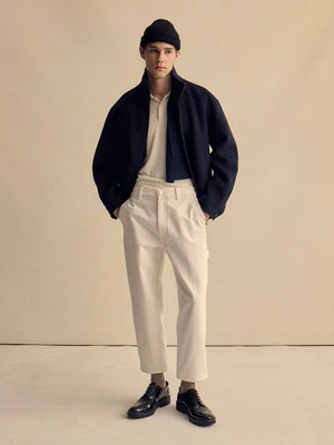 Model wearing a dark jacket, light polo, white trousers, and black beanie standing against a neutral studio backdrop