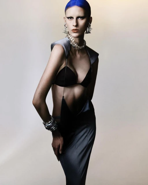 Fashion model with blue hair poses indoors, wearing a bra top, skirt, and futuristic jewelry, against a neutral backdrop in studio lighting.