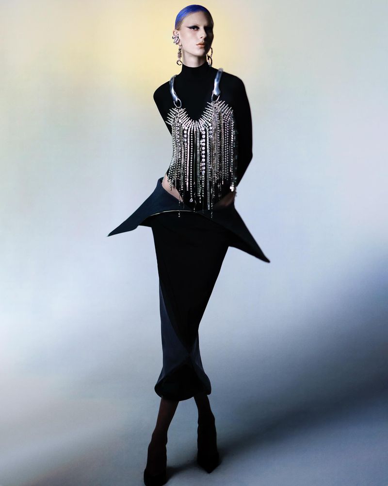 Elegant woman with blue hair poses in a black turtleneck, skirt, and metallic fringe top against a soft gradient background.