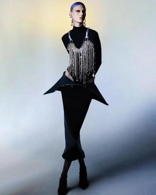 Elegant woman with blue hair poses in a black turtleneck, skirt, and metallic fringe top against a soft gradient background.