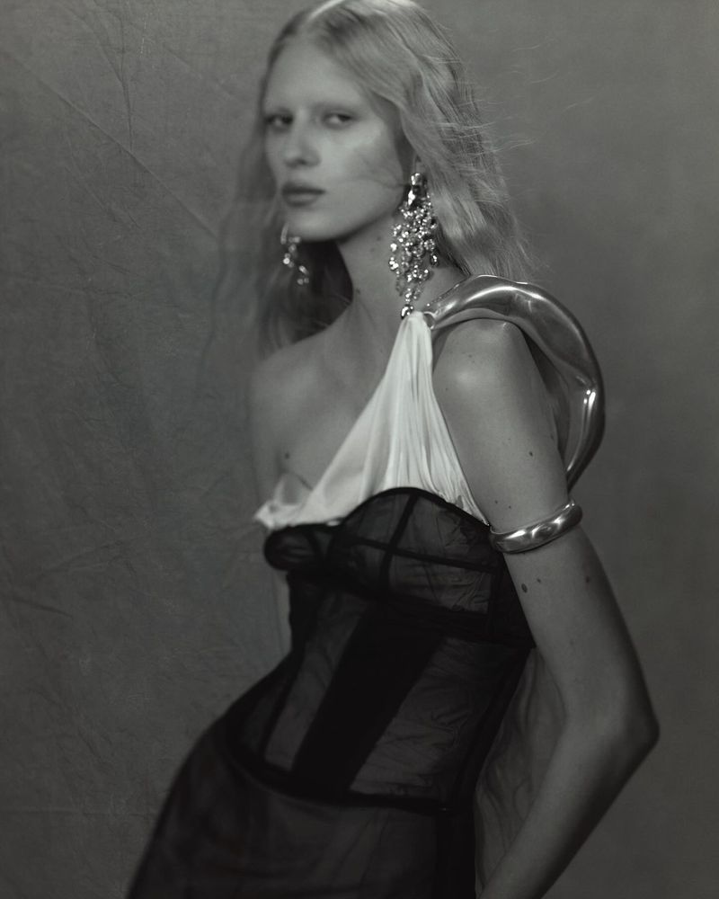 Monochrome portrait of an adult female fashion model with jewelry in a one-shoulder gown against a textured grey backdrop.