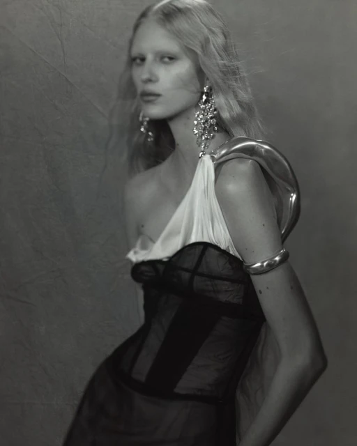 Monochrome portrait of an adult female fashion model with jewelry in a one-shoulder gown against a textured grey backdrop.