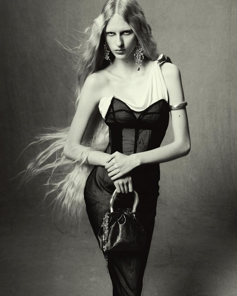 Monochrome portrait of a poised woman with long blonde hair, wearing a one-shoulder dress and holding a silver handbag against a grey backdrop.