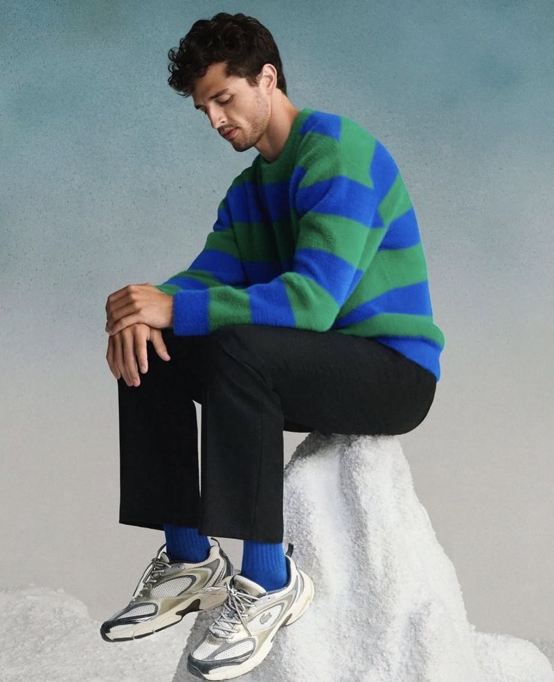 A male model poses sitting on a prop that resembles a snow-covered rock, showcasing a striped green and blue sweater and black pants.