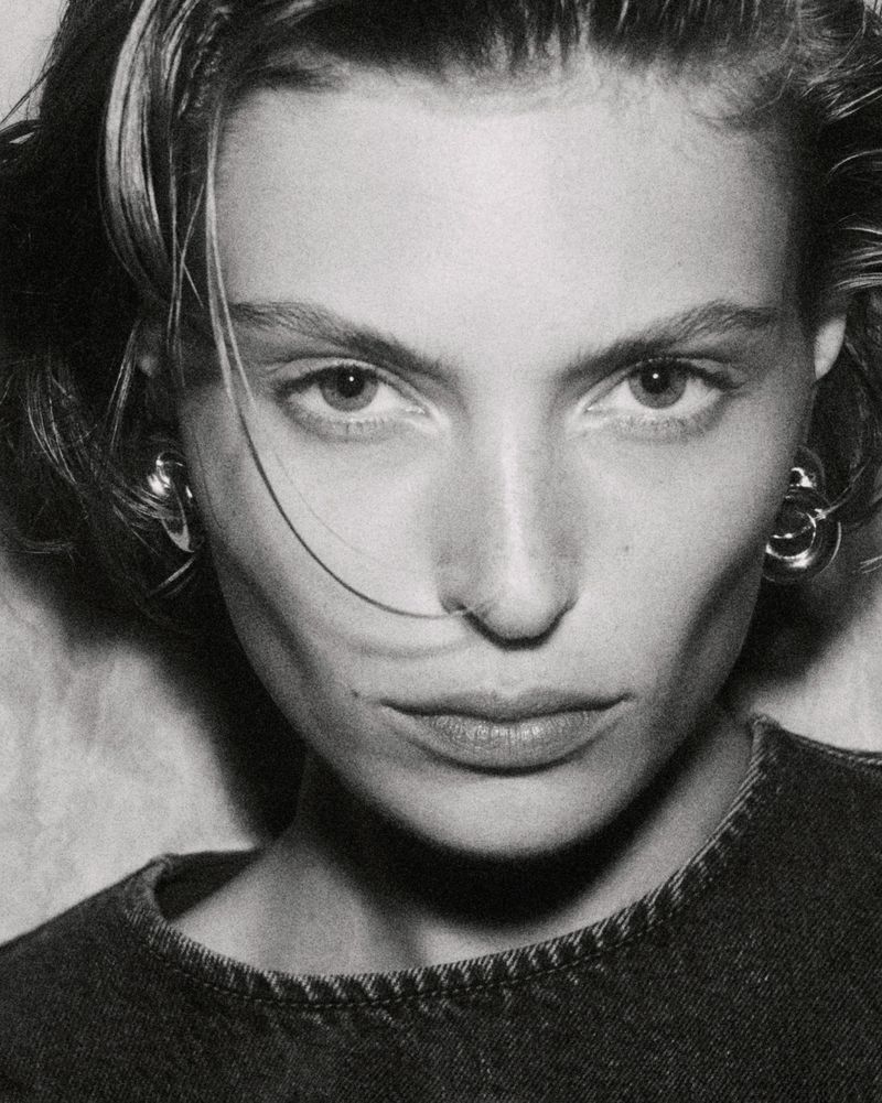 Monochrome portrait of a woman with short hair and round earrings, wearing a dark top against a textured off-white background. Close-up shot.